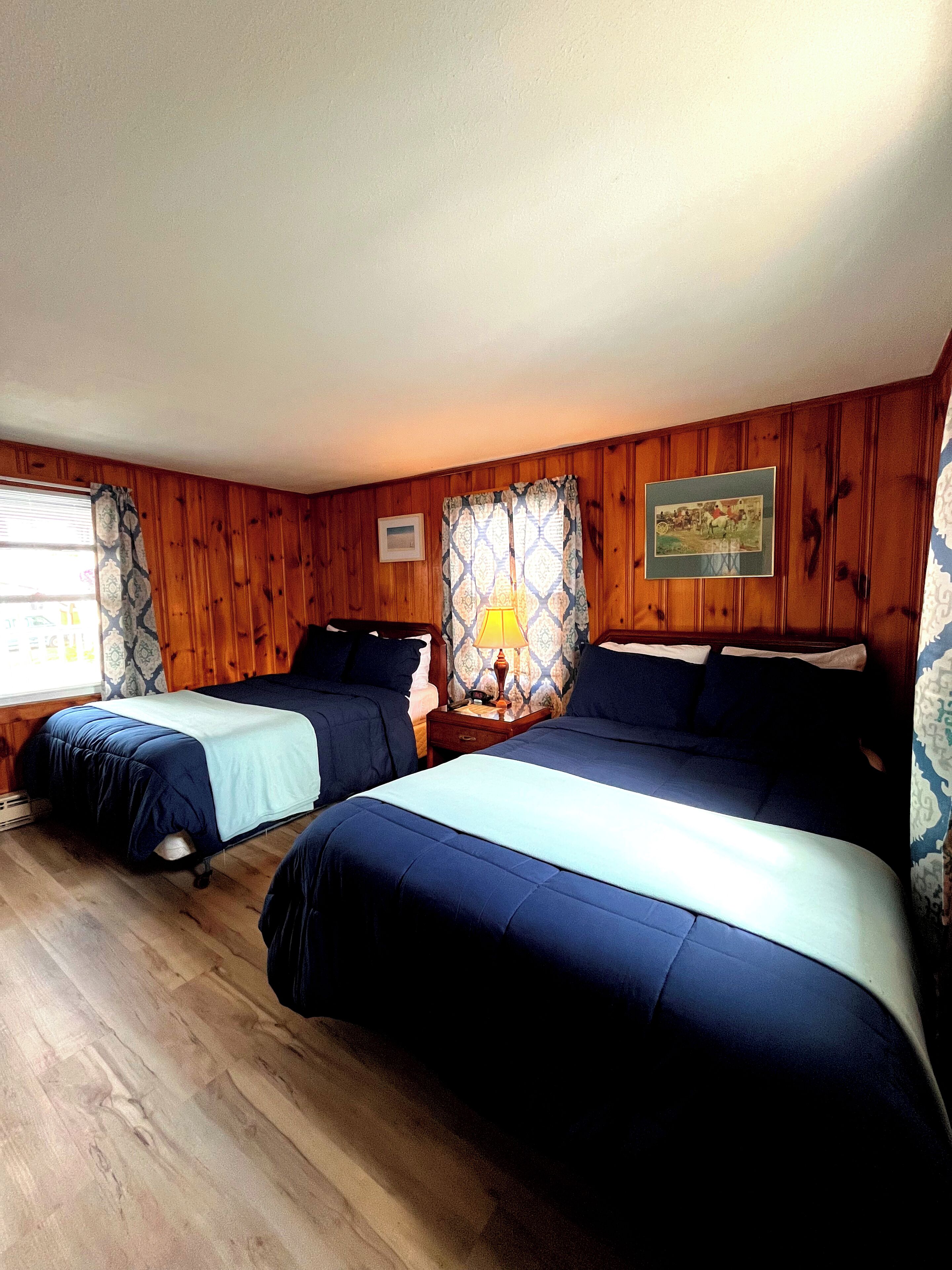 A room with two beds, wooden walls, a window with patterned curtains, a nightstand with a lamp, and a framed picture on the wall.