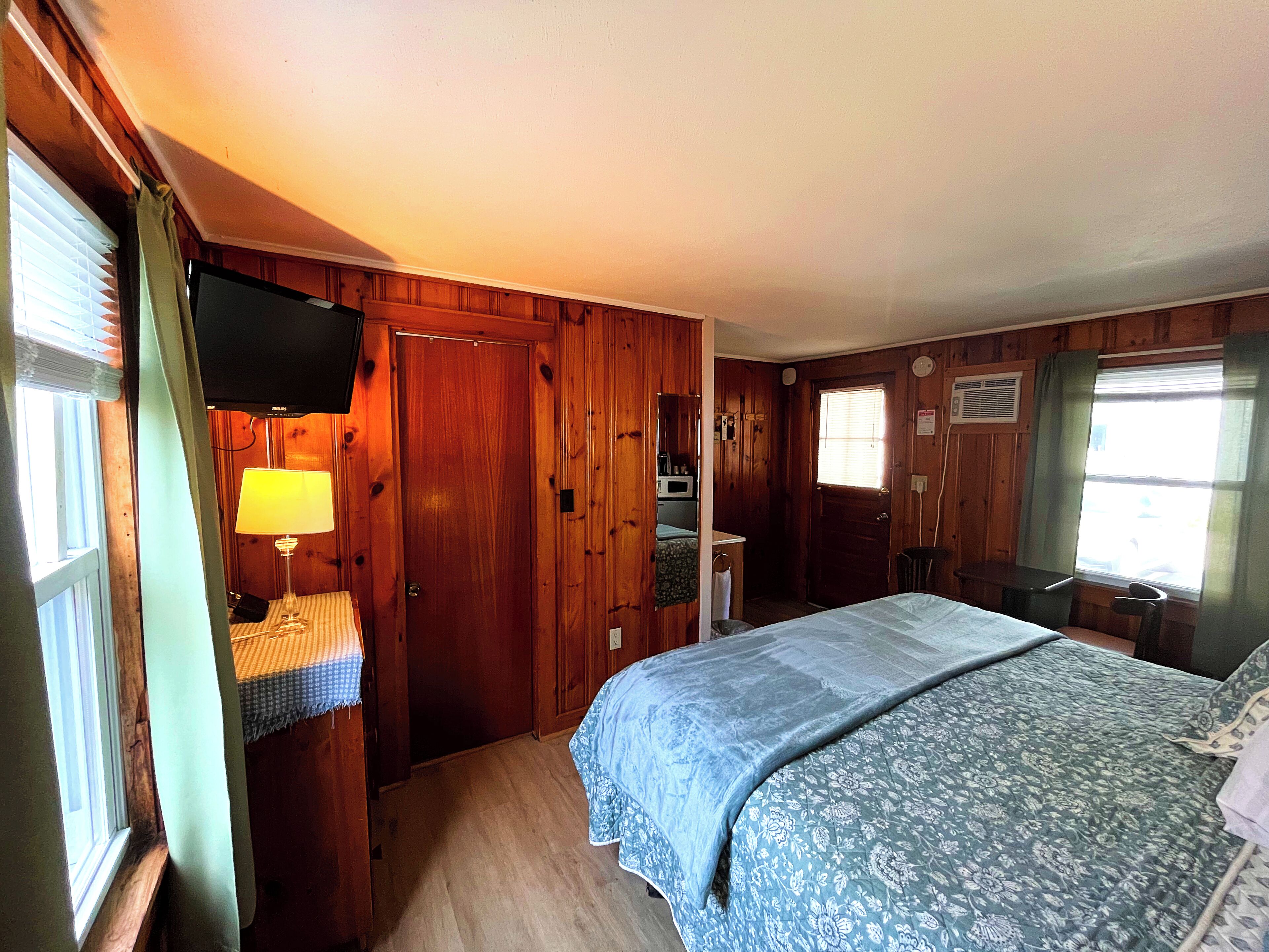 Quarto de madeira com cama, televisão, escrivaninha e janela com cortinas.