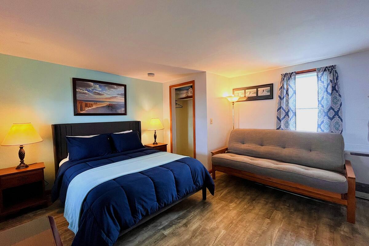 A hotel room with a bed, a sofa, a nightstand with a lamp, a picture on the wall, and a window with curtains.