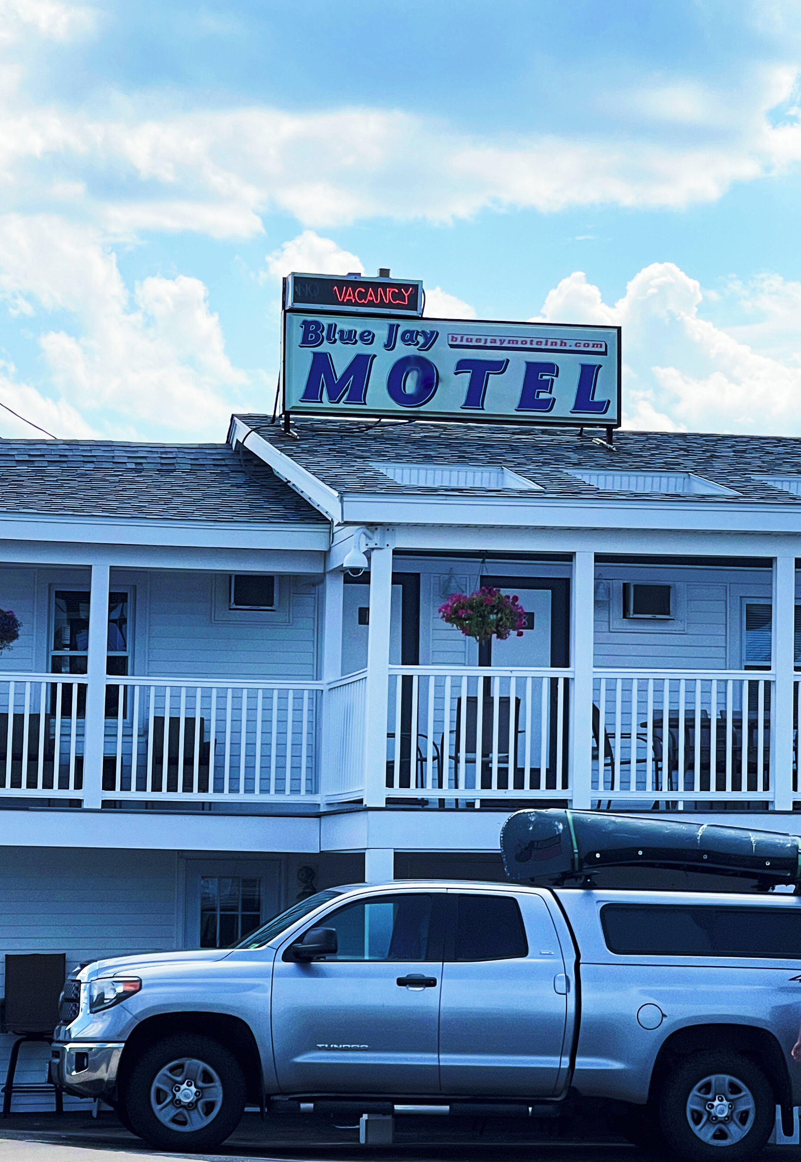 A motel with a 'VACANCY' sign, a porch with a flower basket, and a parked truck.