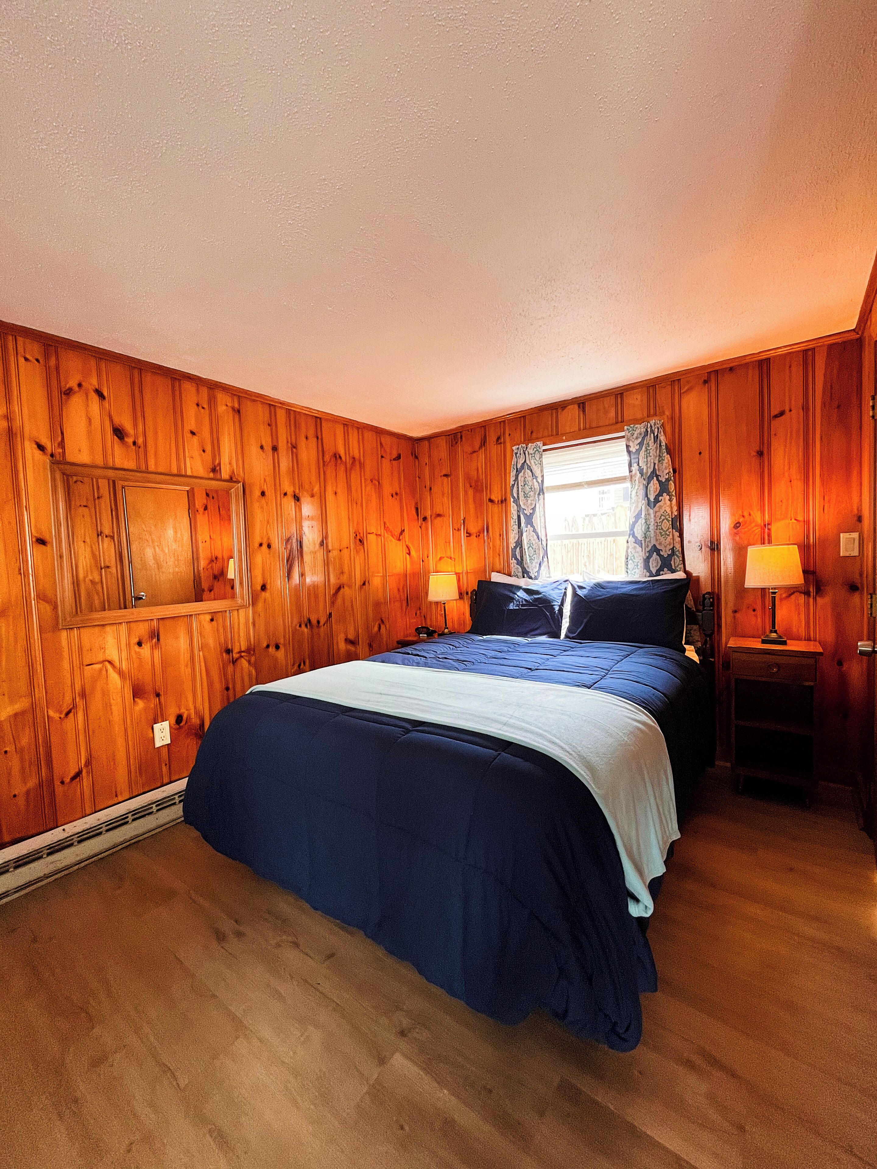 A bedroom with wooden paneling, a bed with blue bedding, two bedside lamps, and a window with patterned curtains.