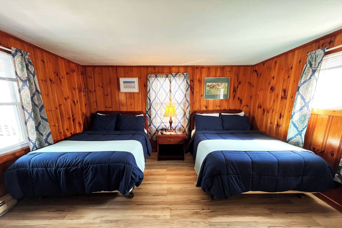 A room with two beds, wooden walls, a nightstand with a lamp, and a window with patterned curtains.