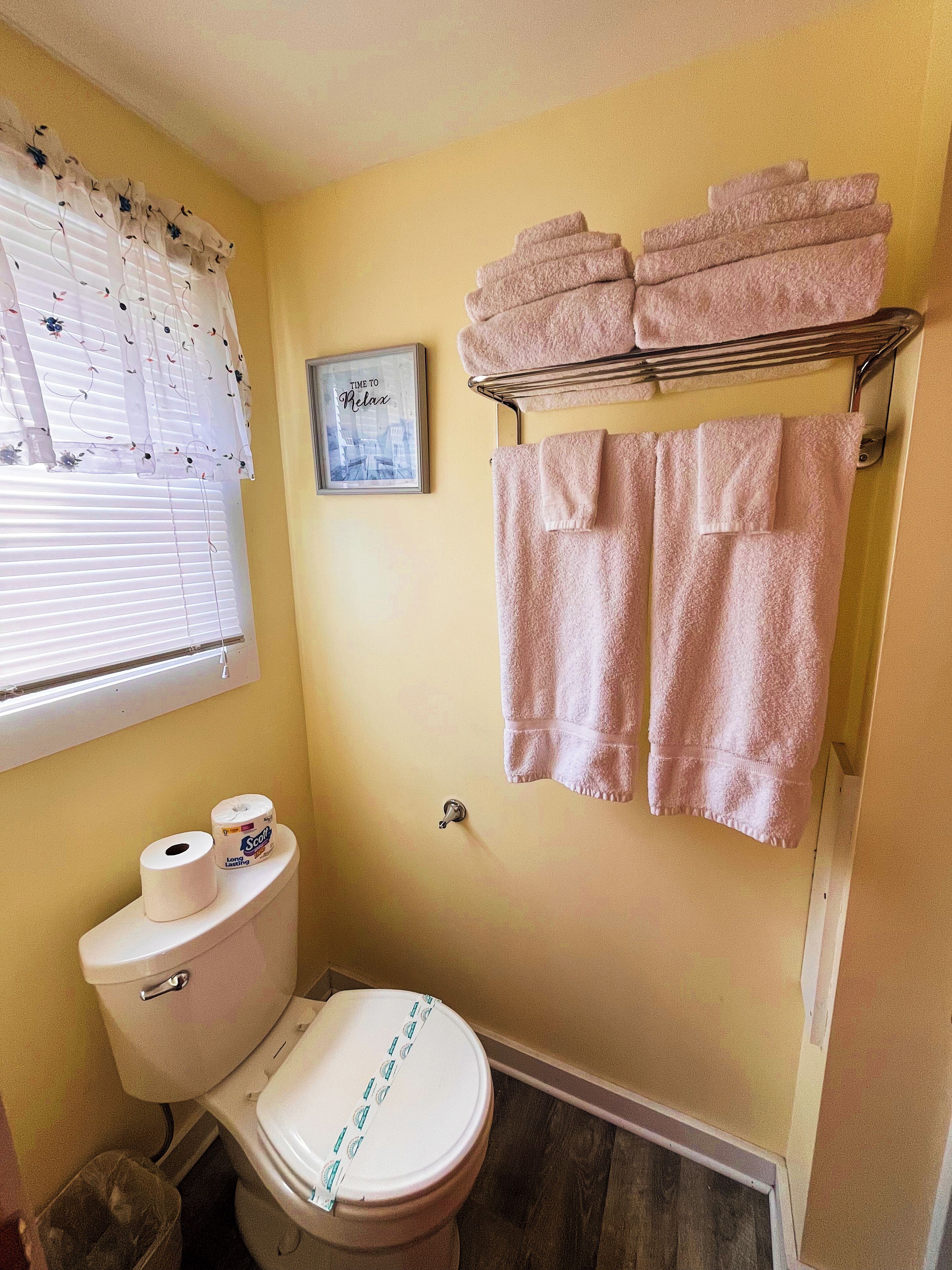 Een badkamer met een toilet, een rol toiletpapier, een handdoekrek met handdoeken en een lijst aan de muur.