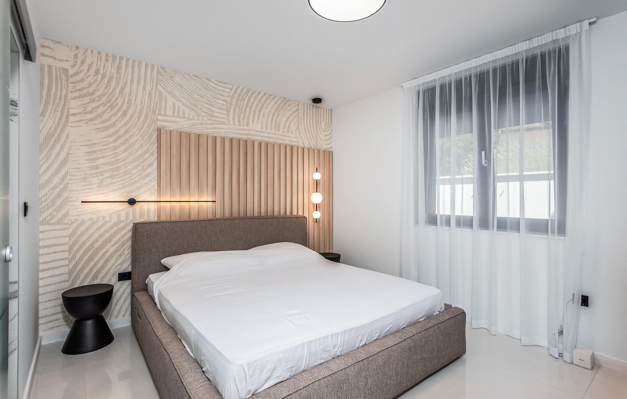 Quarto moderno com cama grande, luminária fixada na parede e cortinas leves.