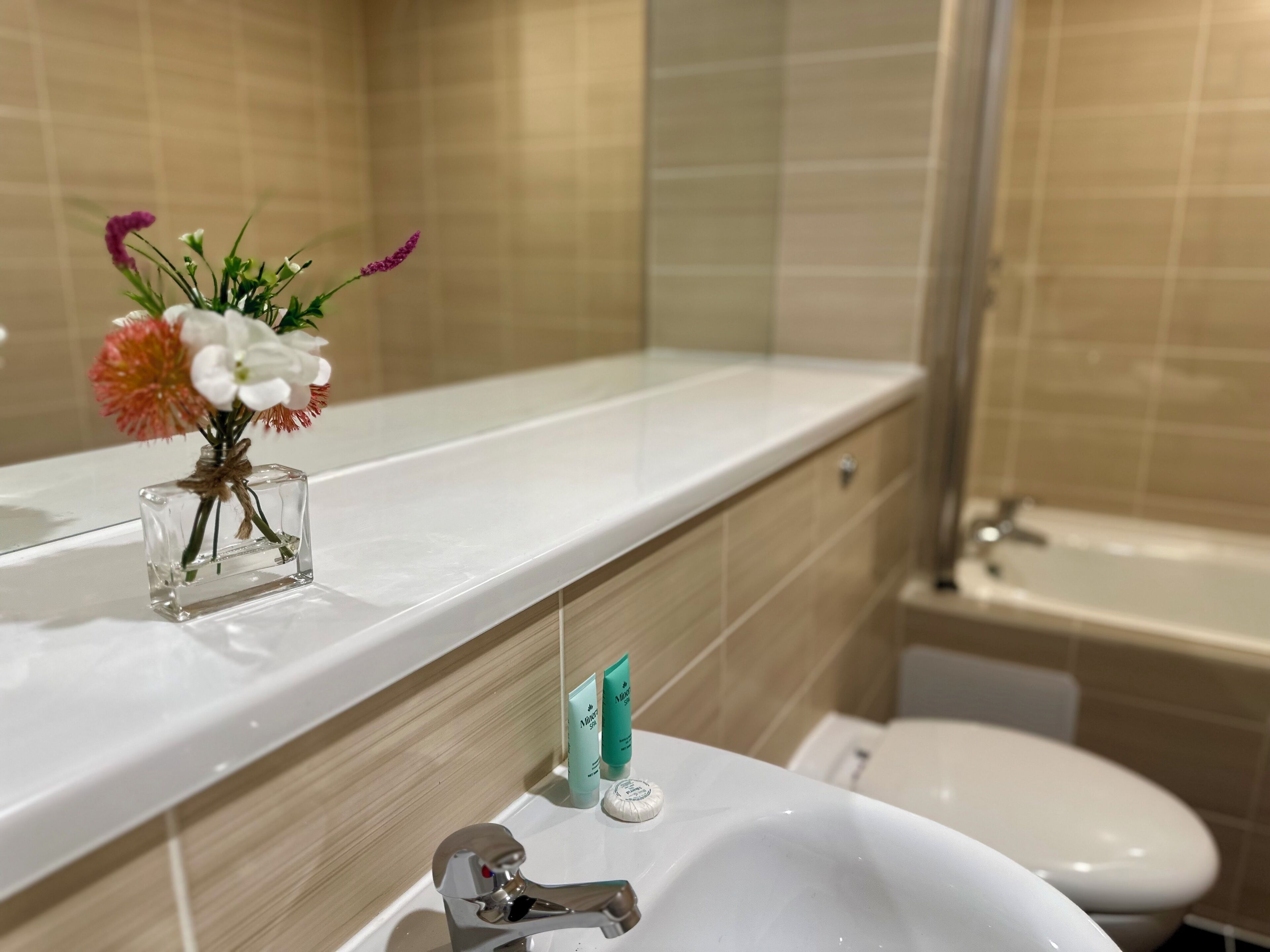 Um banheiro com pia branca, um espelho e um pequeno vaso com flores no balcão.