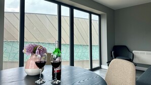 Ein modernes Zimmer mit großem Fenster, einem runden Tisch mit Blumen in einer Vase und zwei Gläsern Rotwein, sowie einem schwarzen Stuhl.