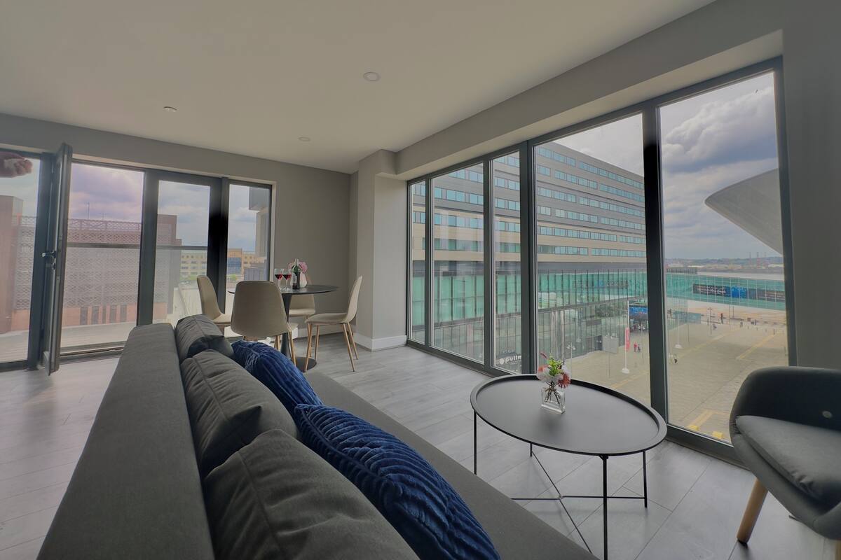 A modern living room with a large sofa, a dining table with chairs, and a small round table with a vase.
