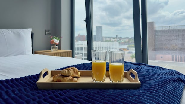 Ein Hotelzimmer mit Bett, einem Tablett mit Croissants und Orangensaft sowie Blick auf die Stadt durch große Fenster.