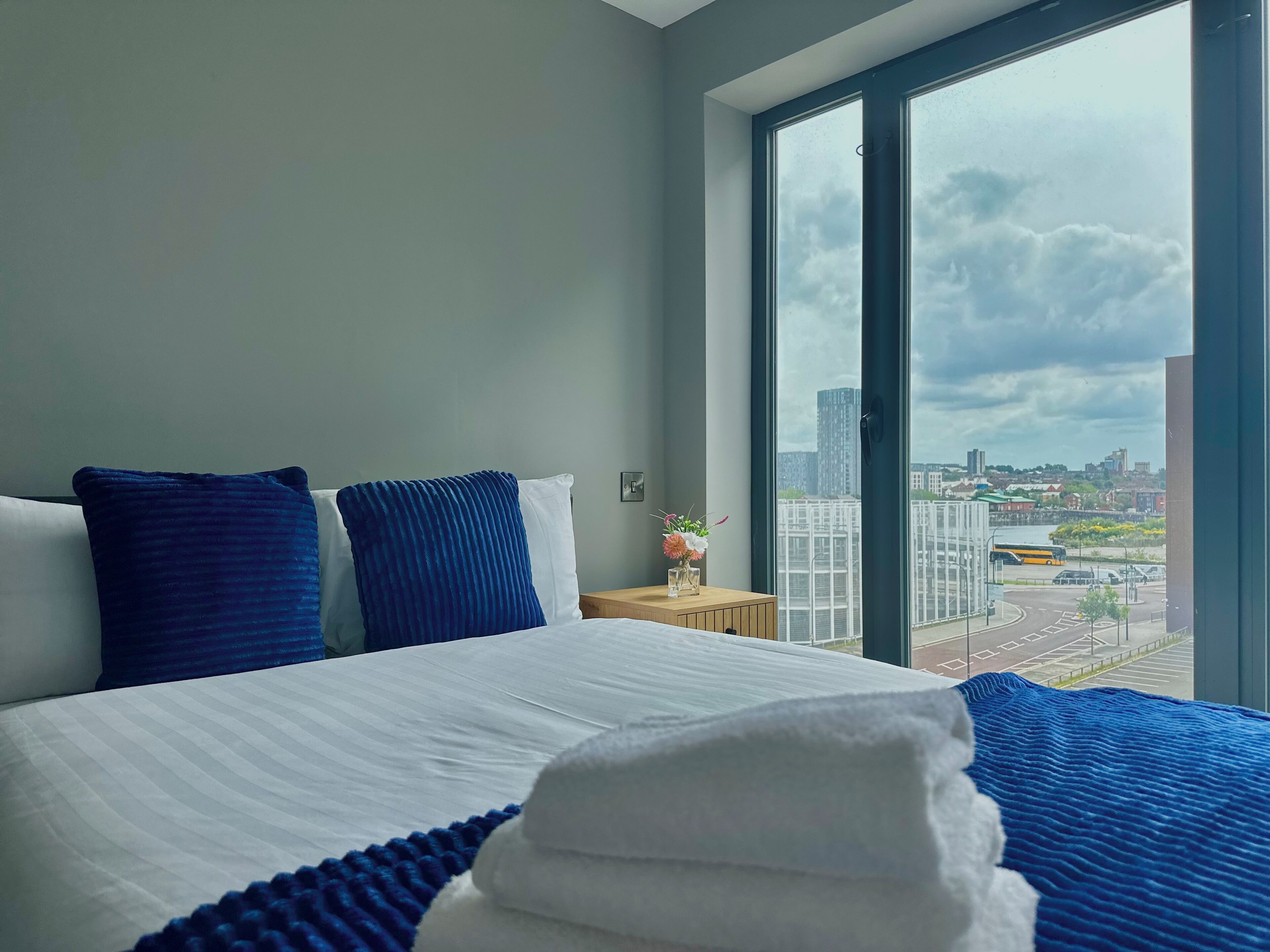 Et hotelværelse med en stor seng, to puder, et natbord med en vase og udsigt over bybilledet gennem et stort vindue.