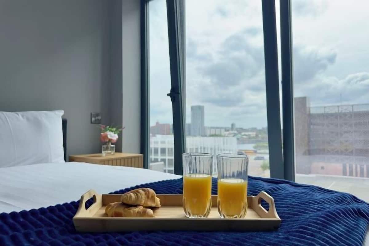 A hotel room with a bed, a tray with croissants and orange juice, and a city view through large windows.