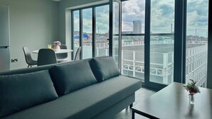 Ein modernes Wohnzimmer mit grauem Sofa, einem kleinen Tisch mit einer Vase und einem Essbereich mit Stühlen und Tisch. Große Fenster bieten Blick auf die Stadtlandschaft.