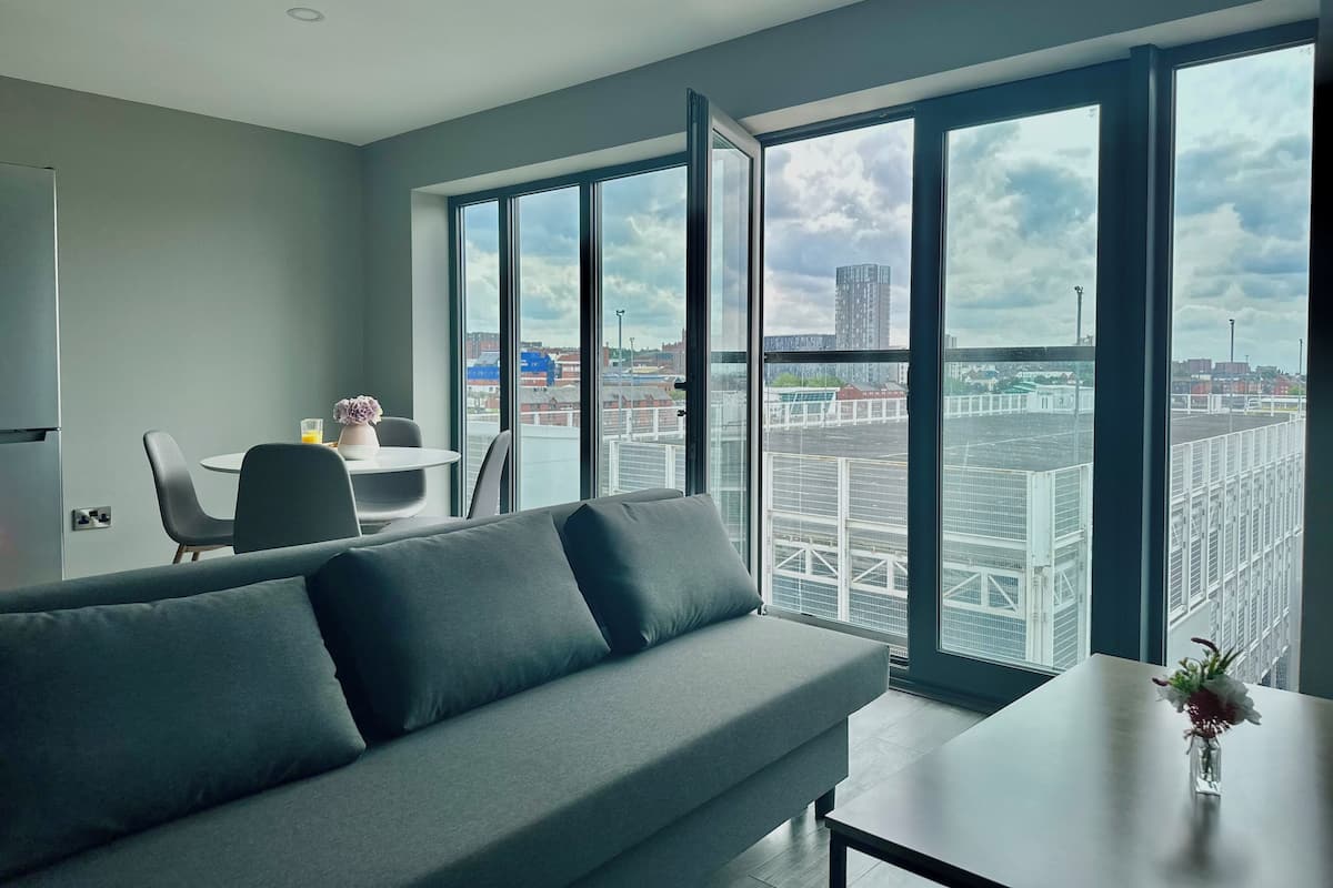 A modern living room with a grey sofa, a small table with a vase, and a dining area with chairs and a table. Large windows offer a view of the cityscape.