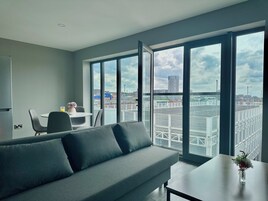 A modern living room with a grey sofa, a small table with a vase, and a dining area with chairs and a table. Large windows offer a view of the cityscape.