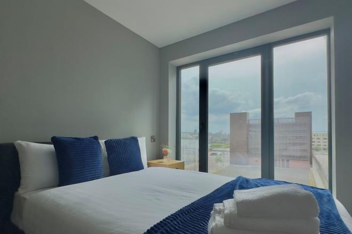 A hotel room with a large bed, a nightstand, and a view of the city through large windows.