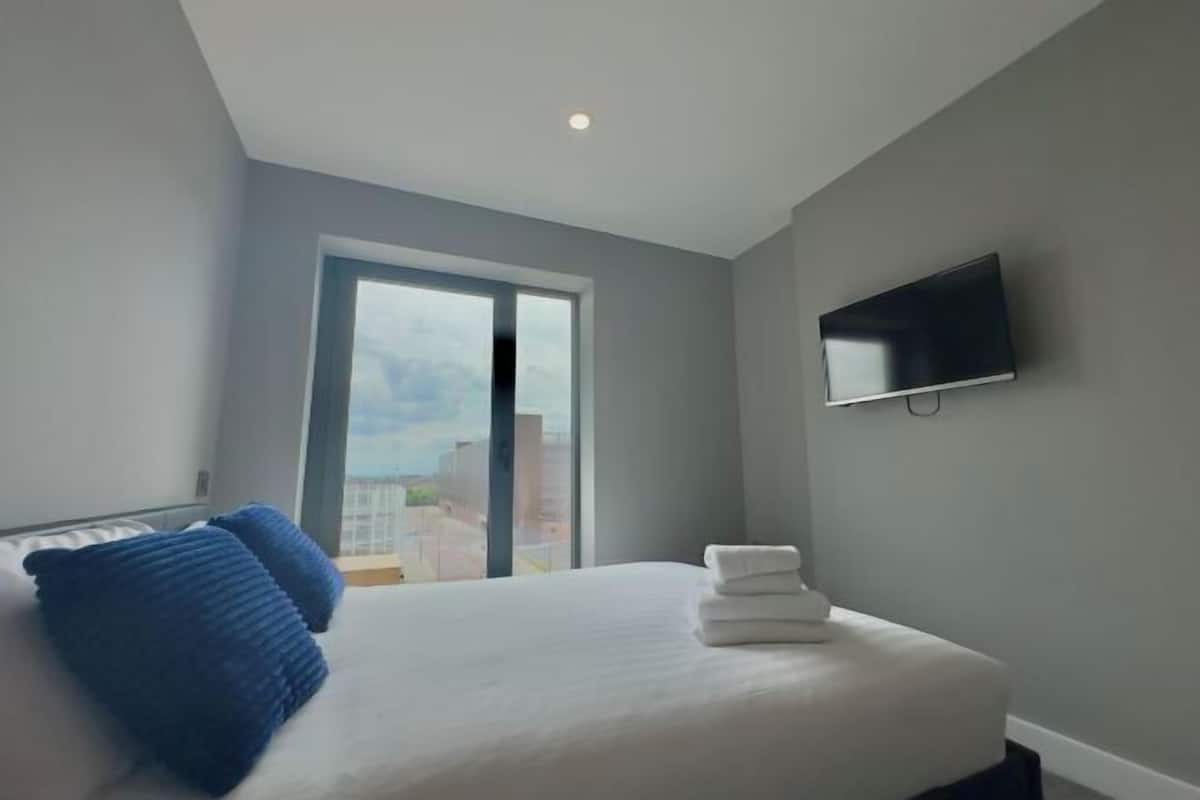 A hotel room with a bed, a TV mounted on the wall, and a window with a view of buildings.