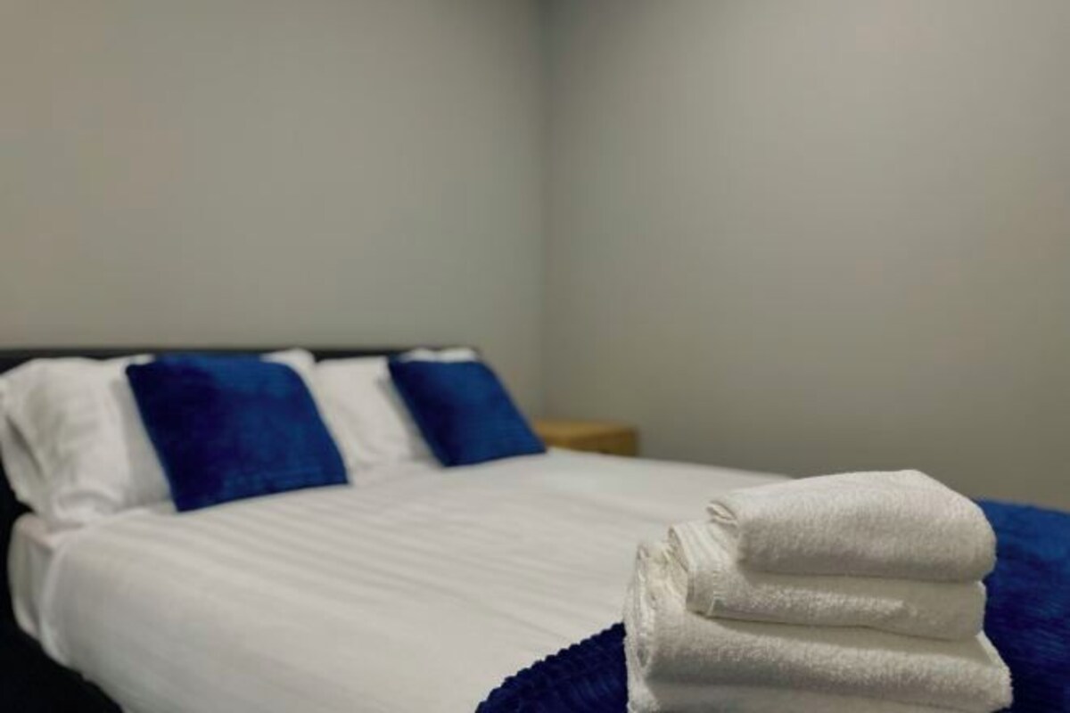 Ein ordentlich bezogenes Bett mit weißer und blauer Bettwäsche, ein gefaltetes weißes Handtuch und ein Nachttisch aus Holz.