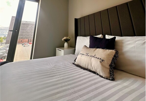 A neatly made bed with striped bedding and a pillow with text, a bedside table with a vase of flowers, and a view of a parking area through a window. - City Centre Albert Dock & Arena Apts (Liverpool)