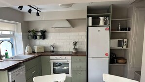 A compact kitchen with a built-in oven, a refrigerator, and a microwave. There are two white chairs around a small dining table.