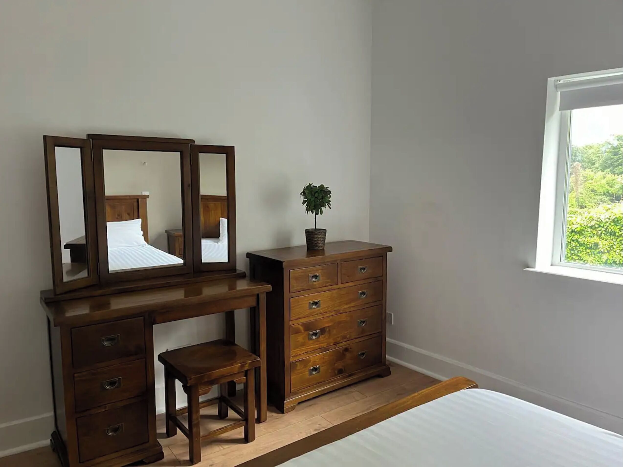 Une chambre à coucher avec une commode en bois, un petit tabouret, un miroir et une fenêtre donnant sur un paysage verdoyant.