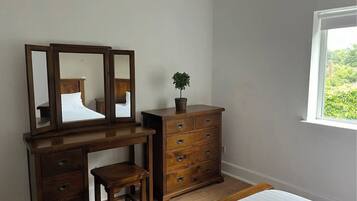 Une chambre à coucher avec une commode en bois, un petit tabouret, un miroir et une fenêtre donnant sur un paysage verdoyant.