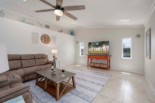 A living room with a brown sectional sofa, a wooden coffee table, a lamp, and a painting of palm trees. - Laguana - Heated Salt Water Pool and Pool Table (Cape Coral)