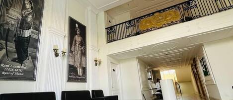 A spacious lobby with a high ceiling, ornate ceiling design, and a grand chandelier. There are black sofas and a white reception desk.