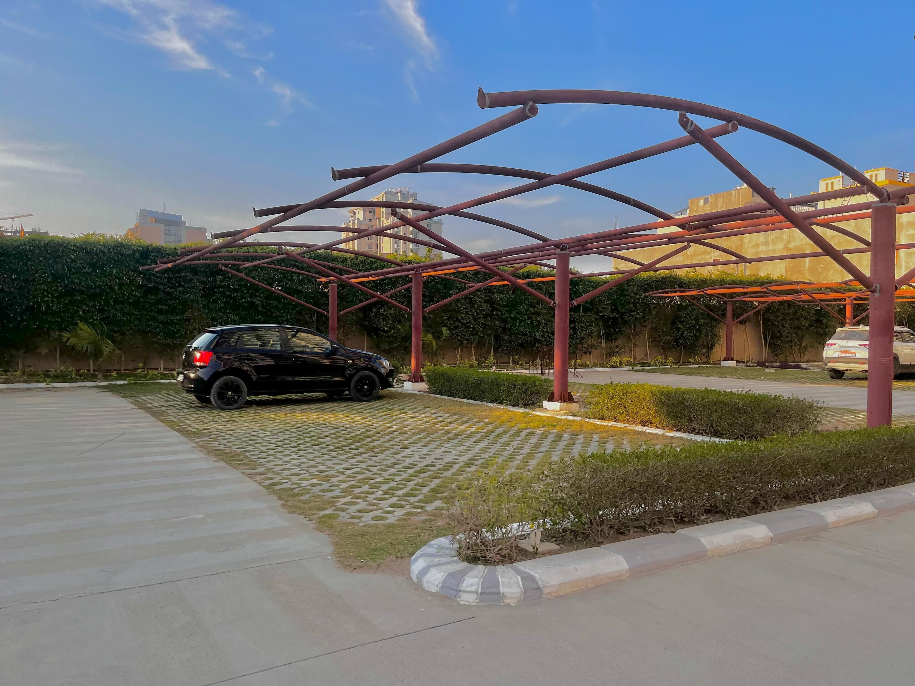 Un’area di parcheggio con un’auto sotto una struttura metallica, circondata da vegetazione e edifici sullo sfondo.