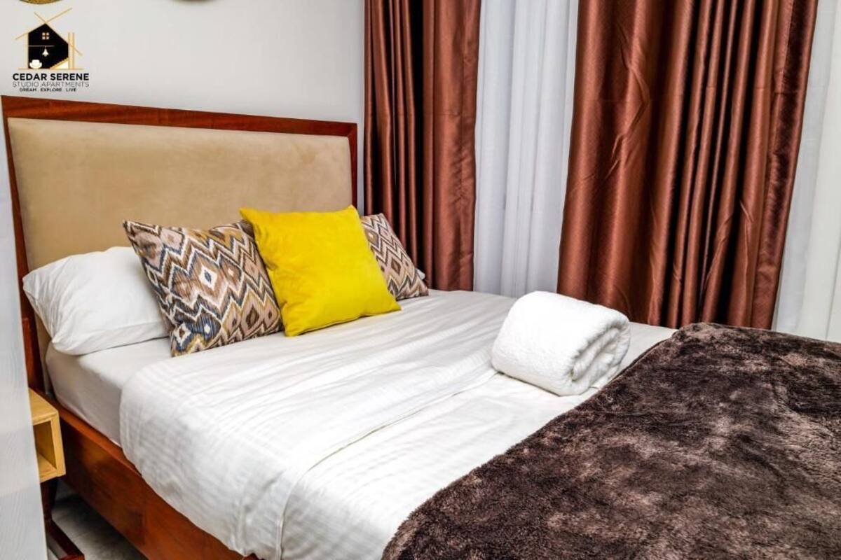 A neatly made bed with a patterned pillow, a yellow pillow, and a brown throw blanket.