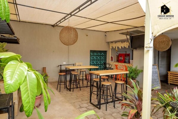 Un espace extérieur accueillant, aménagé avec des tables en bois et des tabourets en métal noir, et agrémenté de différentes plantes en pot.