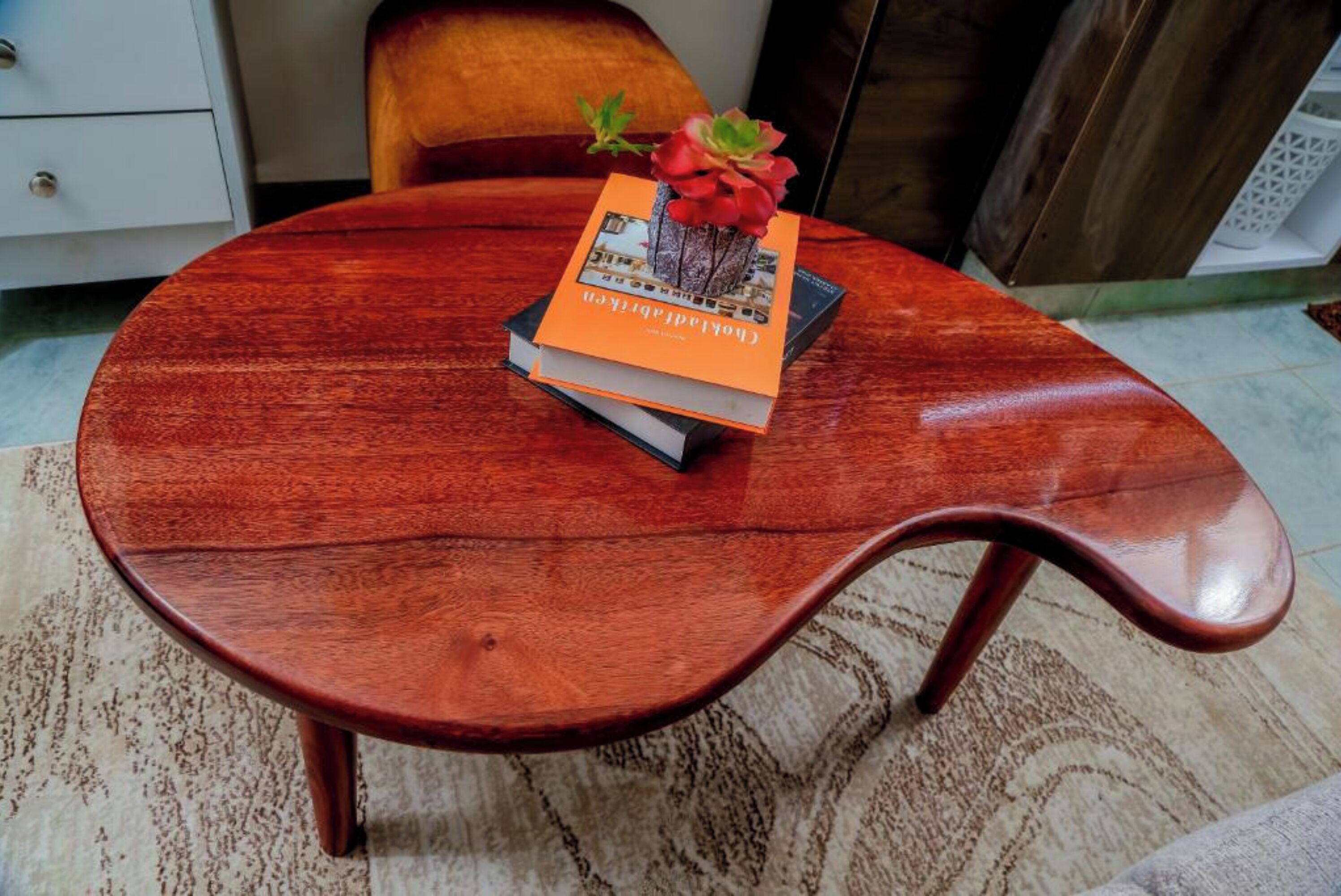 Una mesa redonda de madera con borde curvo, un libro de tapa naranja y una pequeña maceta con flores rojas.