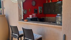 Un espacio de cocina al aire libre con dos taburetes de bar, una pared roja, un reloj y un frasco grande.