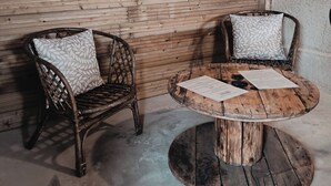 Pared de madera rústica con tres espejos colgantes y un espejo de sol tejido. Hay dos sillas de mimbre con cojines estampados y una mesa redonda de madera con papeles.