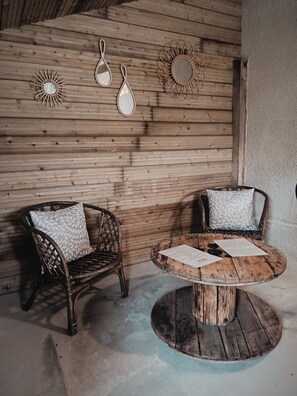 A rustic wooden wall with three wall-mounted mirrors and a woven sun mirror. There are two wicker chairs with patterned cushions and a wooden round table with papers.