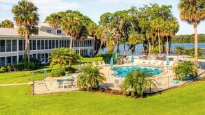 A pool area with lounge chairs and umbrellas, surrounded by palm trees and a building with a view of the water. - Pool Access & Lanai: Riverfront Welaka Condo! (Welaka)