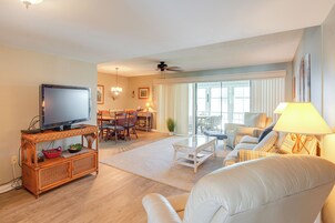 Una sala de estar con televisor, muebles de mimbre, una zona de comedor y un ventanal grande.