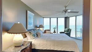 A bedroom with a large bed, two bedside lamps, a ceiling fan, a chair, and a window with blinds. - Edgewater Tower II #803 | Incredible Views + On-Site Pool & More! (Panama City Beach)