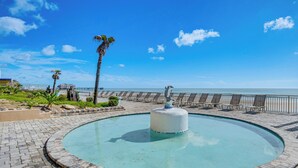 A circular pool with a central sculpture, surrounded by a paved walkway and beach chairs, overlooking a beach and ocean. - Cozy Daytona Beach Condo - Steps to Ocean! (Daytona Beach)