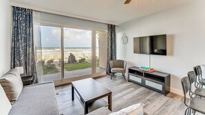 A modern living room with a large sliding glass door to the outdoors, a flat-screen TV mounted on the wall, a sofa, a coffee table, and a TV stand with a few items on it. - Cozy Daytona Beach Condo - Steps to Ocean! (Daytona Beach)