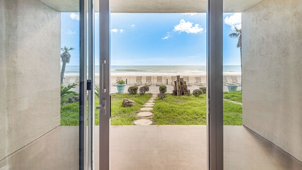 A view from a room with a glass door leading to a balcony with a stone pathway, palm trees, and a beach in the background. - Cozy Daytona Beach Condo - Steps to Ocean! (Daytona Beach)