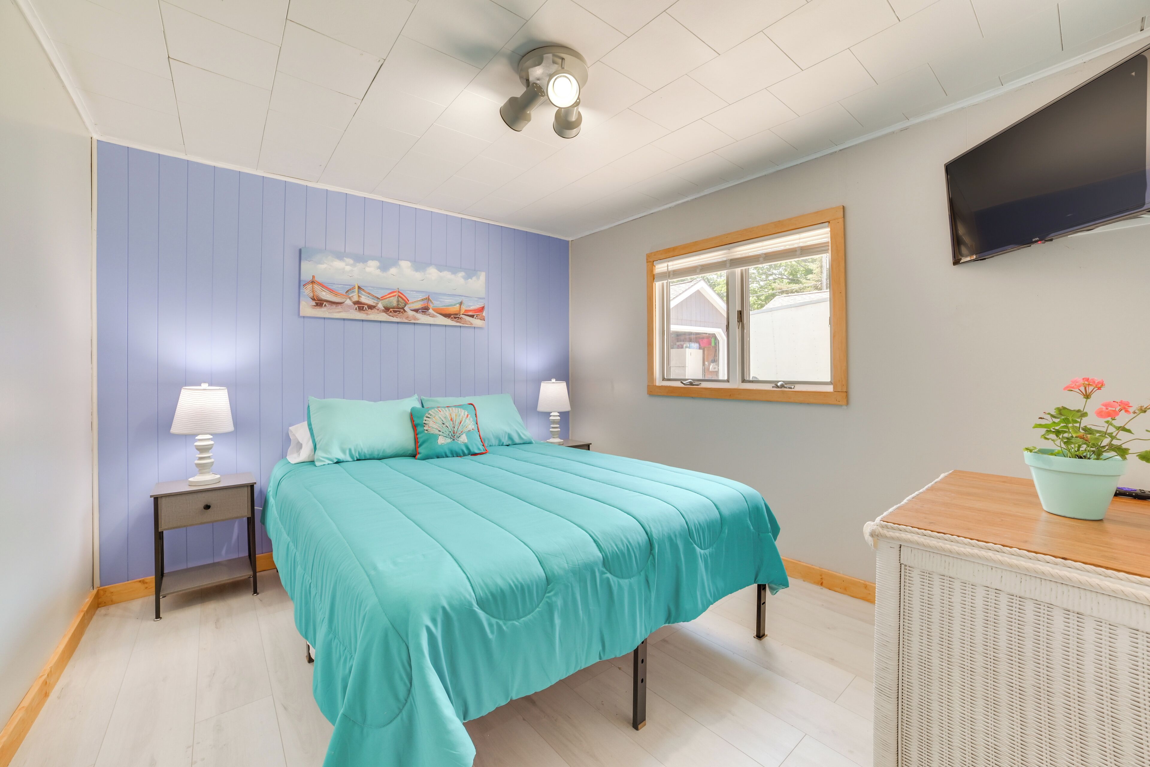 A bedroom with a bed, nightstands, a wall-mounted TV, a window, and a potted plant.