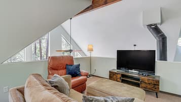 A loft-style living room with a sloped ceiling, a large window, a leather armchair, a sofa, a TV on a wooden stand, and a floor lamp.