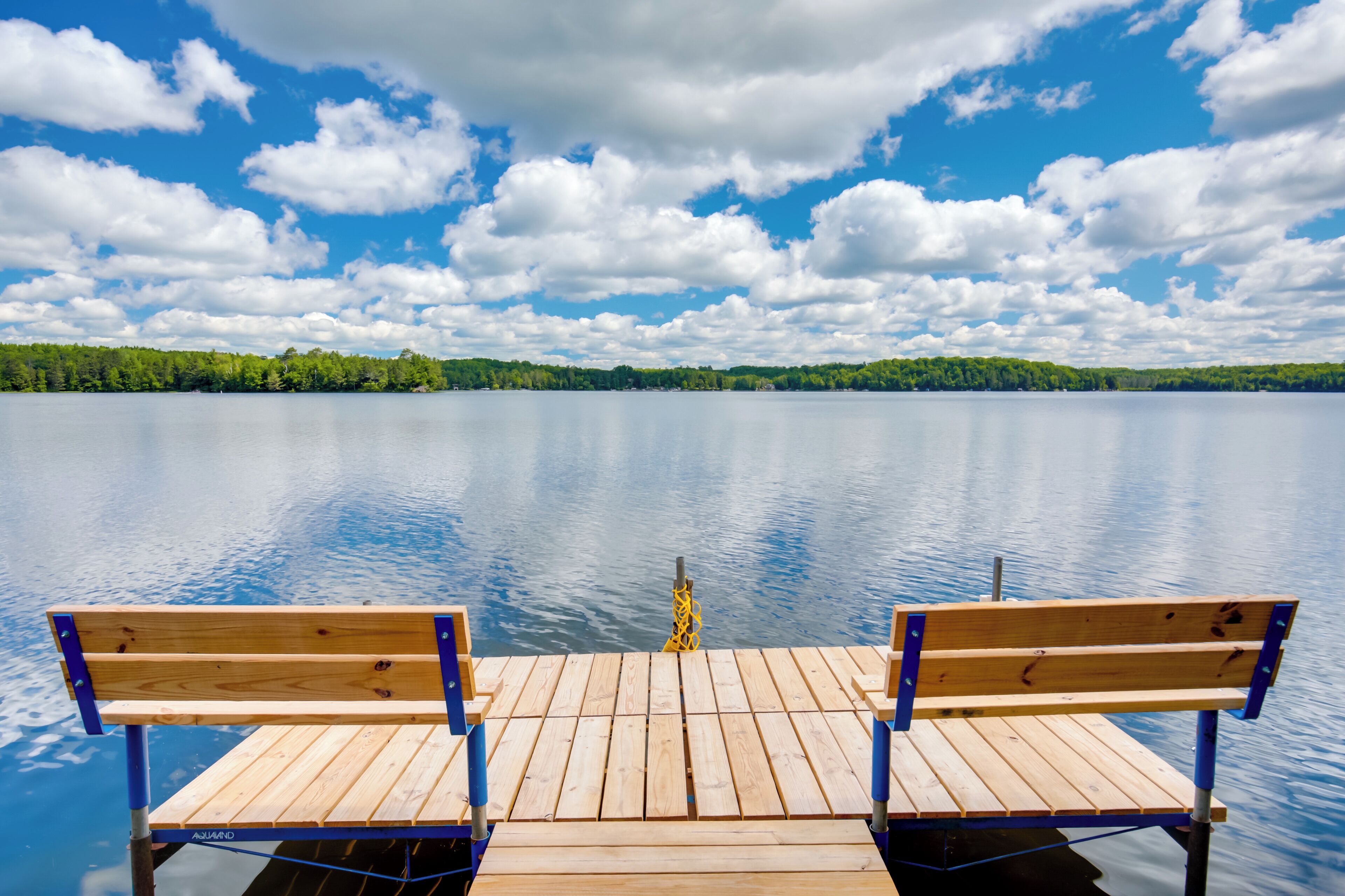 A wooden dock with benches extending over calm water, surrounded by a forested shoreline under a partly cloudy sky.