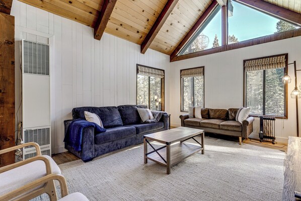 A spacious living room with a high ceiling, large windows, a blue sofa, a brown sofa, and a wooden coffee table. - Bright Truckee Cabin Close to Skiing & Donner Lake (Truckee)