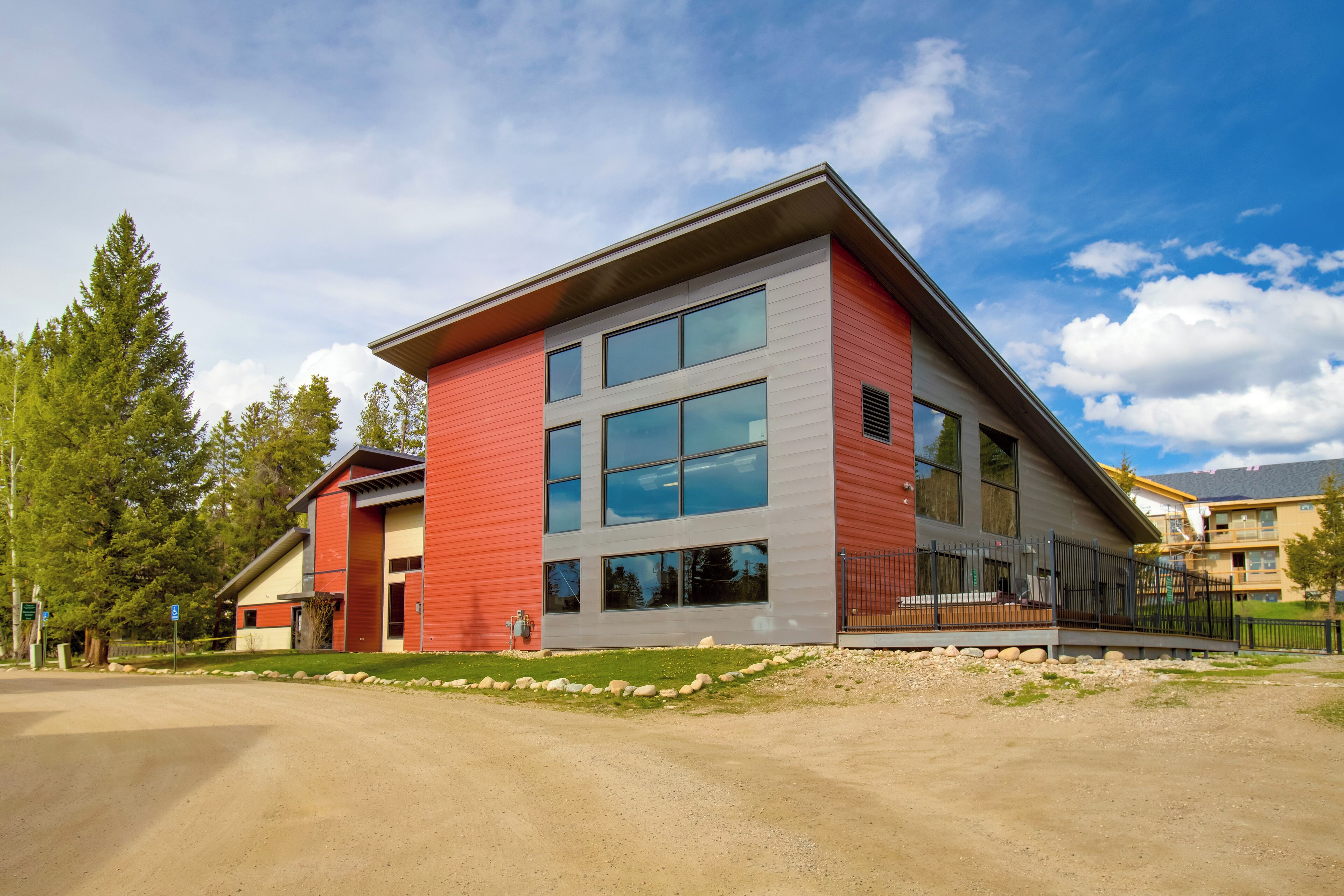 A modern building with a red and grey facade, large windows, and a paved driveway.