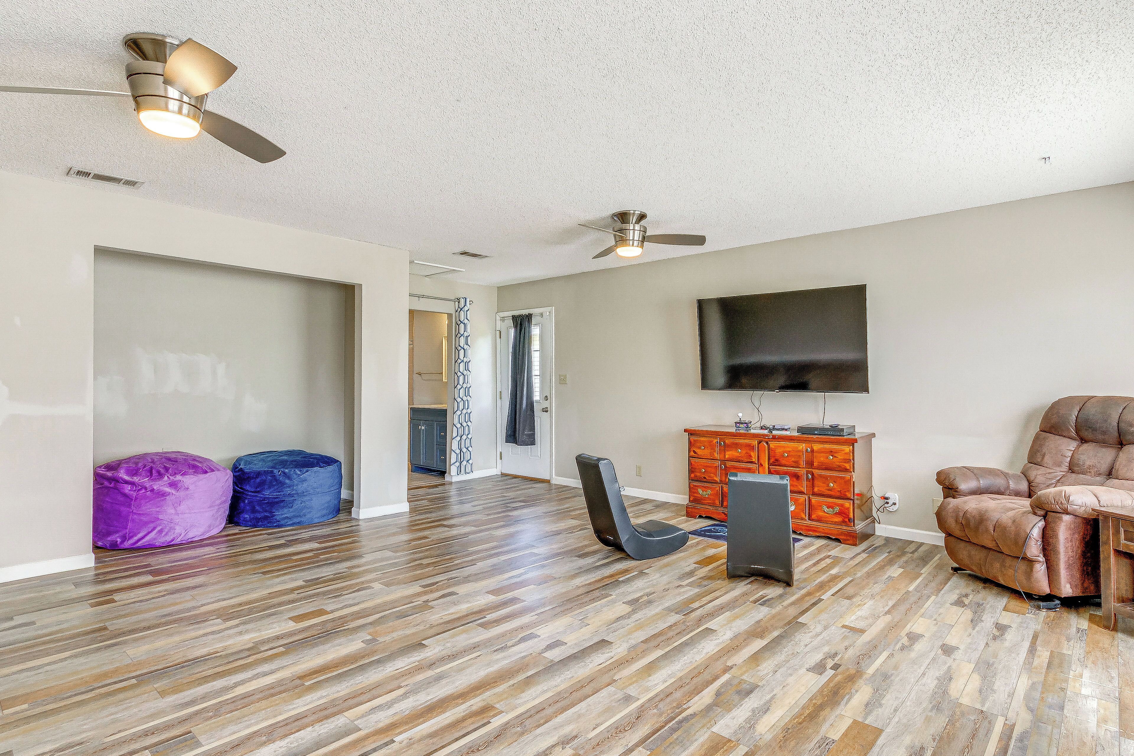 A spacious living area with hardwood flooring, a ceiling fan, a leather recliner, a flat-screen TV, and a wooden cabinet.