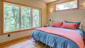 Un dormitorio con un ventanal grande, una cama con ropa de cama azul y roja, y una mesita de noche con una lámpara.