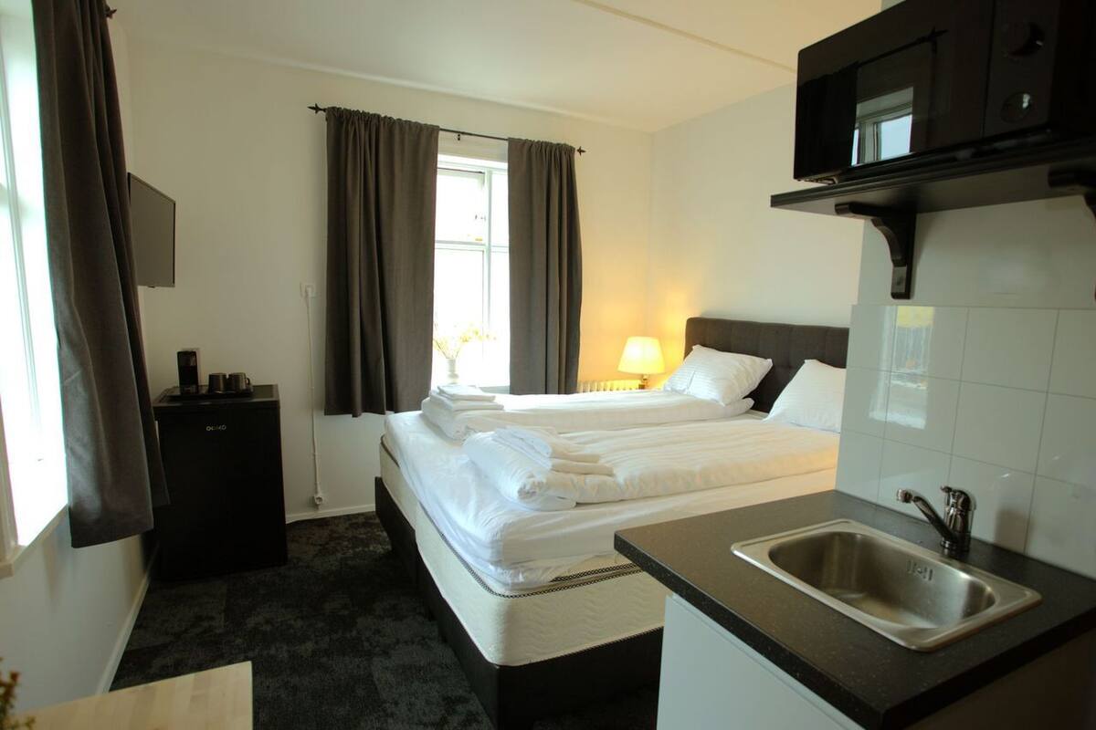 Habitación de hotel con cama, mesita de noche, lámpara, televisión y zona de cocina con lavabo.