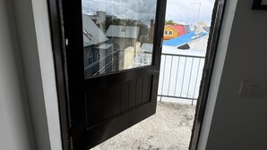 Vista a través de una puerta entreabierta, mostrando un balcón con barandilla metálica y un edificio con tejado rojo al fondo.