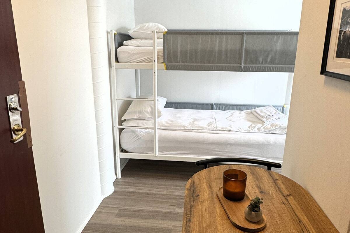 Una habitación pequeña con una cama litera, una mesa de madera con una taza y una planta pequeña, y una puerta con pomo.