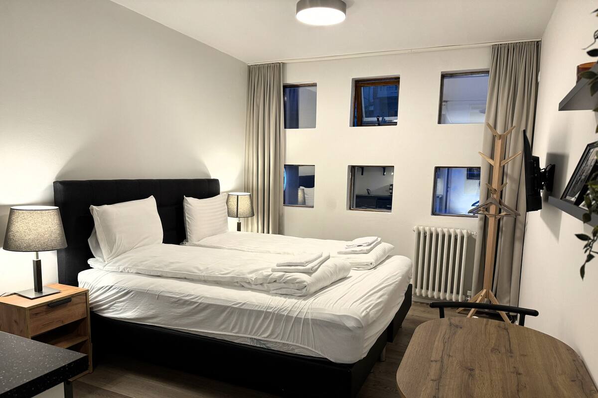 Habitación de hotel con una cama, mesitas de noche, una silla de madera y vistas al exterior a través de ventanas.